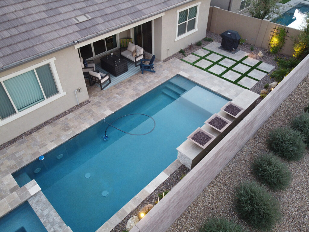 Overhead view of an Arizona backyard with a sleek rectangular lap pool lined by travertine deck and narrow planters, showing artificial turf-grid patio and desert landscaping, built by Phoenix pool builders.