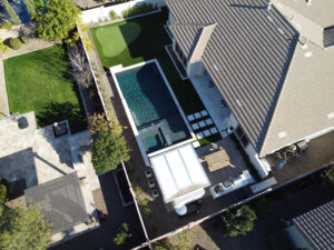 Thumbnail of http://Blurred%20aerial%20view%20of%20an%20Arizona%20property%20showing%20the%20house%20roof,%20rectangular%20lap%20pool,%20travertine%20deck%20and%20green%20lawn%20captured%20by%20Phoenix%20pool%20builders.
