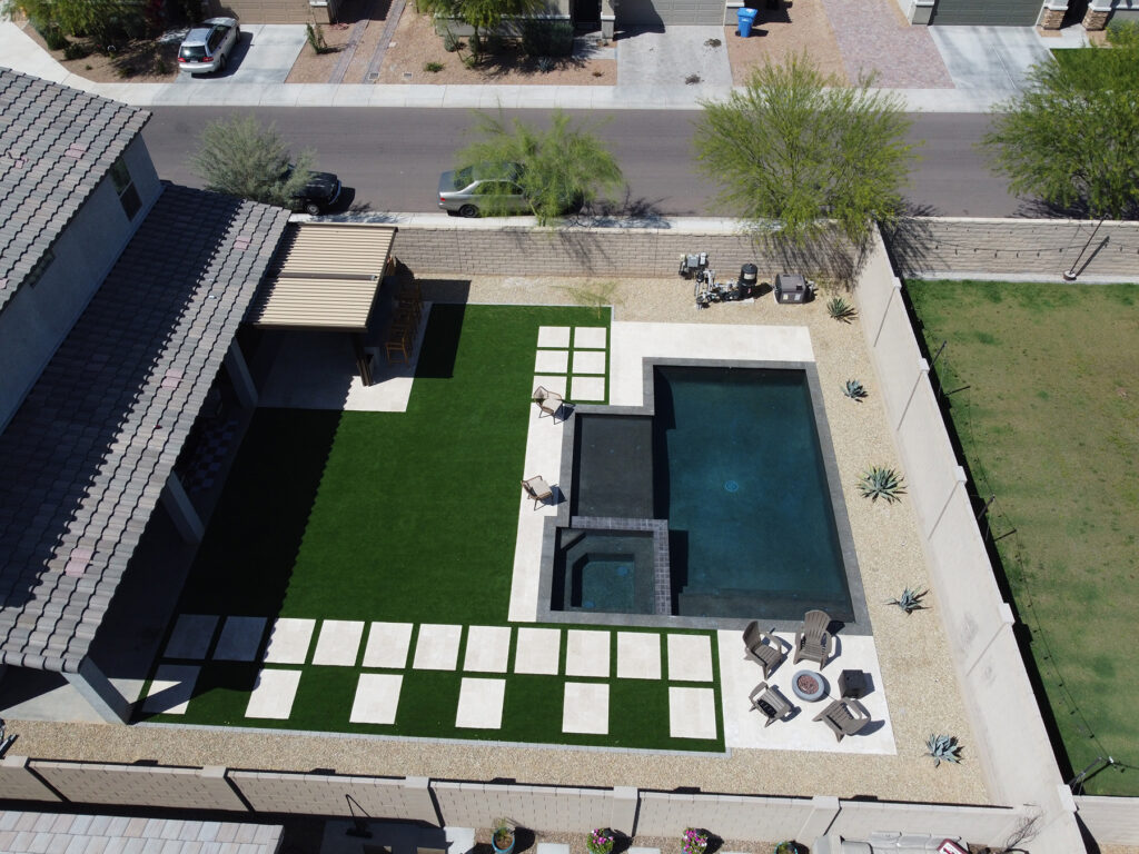 Aerial view of an Arizona backyard featuring a dark rectangular pool with spa, large artificial turf lawn with stepping‑stone pathways, a shaded pergola and gravel landscaping designed by Phoenix pool builders