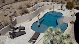 Elevated view of a modern Arizona backyard with a rectangular turquoise pool featuring an attached square spa, geometric paver patio with planters and outdoor seating area, adjacent to a pool house and desert landscaping, built by Phoenix pool builders.