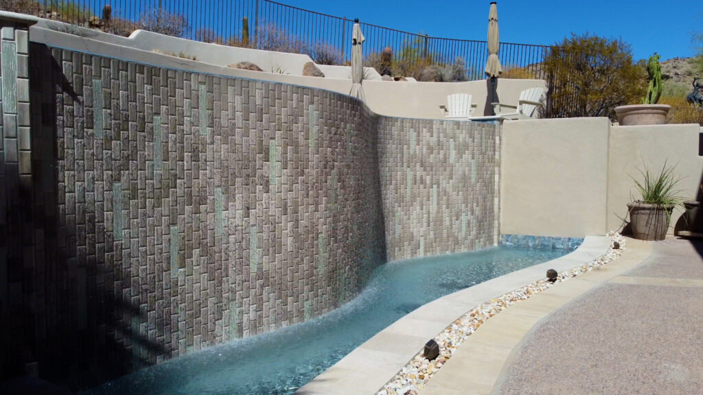 Elevated view of a modern Arizona backyard with a rectangular turquoise pool featuring an attached square spa, geometric paver patio with planters and outdoor seating area, adjacent to a pool house and desert landscaping, built by Phoenix pool builders.