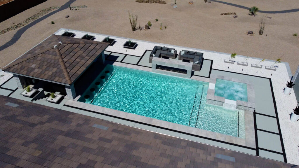 Aerial view of a stylish Arizona backyard featuring a large rectangular turquoise swimming pool with an attached raised spa and shallow tanning ledge, surrounded by travertine deck and a covered poolside bar, overlooking desert landscaping, built by Phoenix pool contractors.