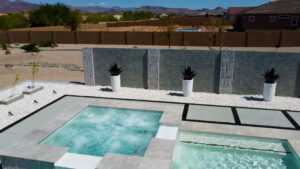 Close-up view of an upscale Arizona backyard featuring a raised square spa with turquoise water adjacent to a rectangular pool, surrounded by modern concrete walls with built-in spillway water features and large potted agave plants, with desert landscape in the background, built by Phoenix pool builders.