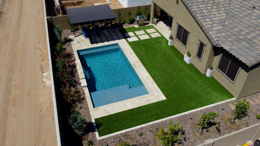 Drone view of a modern Arizona backyard showcasing a dark blue rectangular swimming pool with attached spa, travertine pool deck, pergola shade structure, artificial turf lawn and desert landscaping, built by Phoenix pool builders.